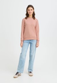 Jasnoróżowa bluza z okrągłym dekoltem, ściągaczami przy mankietach i dole. Dopasowana do jasnoniebieskich szerokich spodni dżinsowych i beżowych butów z ciemnymi sznurowadłami.