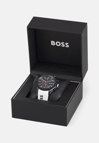 Black watch with a black dial and silver hardware, featuring red accents. Displayed on a cushion inside a black box with "BOSS" branding.