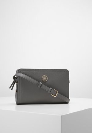 Sac bandoulière rectangulaire en cuir gris foncé avec bandoulière réglable et emblème rond en or du logo à l'avant, posé sur une surface blanche.