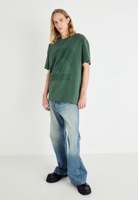 Camiseta de algodón verde con logo, de corte holgado. Combinada con jeans de pierna ancha de color azul claro y zapatos negros de suela gruesa, de pie sobre un fondo sencillo.