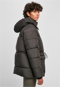 Puffer jacket in dark gray with a hood, featuring a cinched waist, elastic cuffs, and a front pocket with drawstring detail.