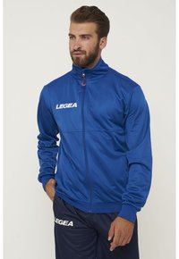 Giacca blu con zip realizzata in tessuto morbido, con colletto alto, tasche laterali e un logo sul petto. Abbinata a pantaloni blu navy coordinati.