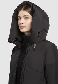 Veste rembourrée noire avec une grande capuche structurée. Présente une fermeture éclair et des cordons de serrage pour réglage. Extérieur lisse et brillant.