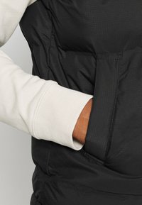 Gilet imbottito nero con superficie testurizzata, caratterizzato da una manica di colore crema visibile al polsino e una tasca laterale aperta.