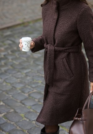 Person iført en brun uldfrakke, der holder en hvid kaffekop med et sort abstrakt ansigtsdesign og en brun læderhåndtaske.