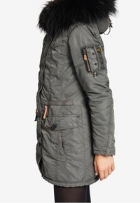 Zijaanzicht van een persoon die een grijze parka met een bont-gevoerde capuchon draagt, met meerdere zakken en ritsdetails op de bovenste mouw.