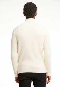 Suéter de punto color crema claro con cuello alto, textura de canalé y mangas largas. Combinado con pantalones negros, mostrando la vista posterior.