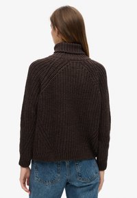 Brązowy sweter z dzianiny, z wysokim kołnierzem, prążkowaną teksturą i rękawami raglanowymi, w zestawie z niebieskimi dżinsami o wysokim stanie.