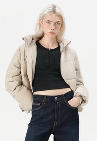 Beige puhvisjakker, millel on kapuuts ja tikanditunne. Must pikkade varrukatega topp all ja tume sinine teksapüksid. Lihtsad nööpide detailid topil.