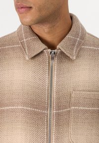 Jachetă cu fermoar, de culoare maro, cu model houndstooth, guler cu guler, buzunar frontal și accente de cusături albe.  Material texturat moale.