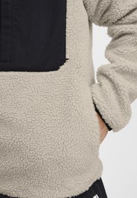 Light beige fleece pullover with a navy panel, textured surface, loose fit, and a front pocket detail. Cuffs are slightly gathered.