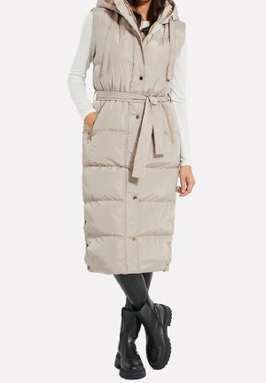 Femme portant un long manteau beige matelassé sans manches avec une ceinture nouée, un haut blanc à manches longues, des leggings noirs et des bottines noires.