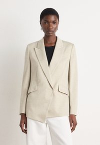 Beige bleiseri, jolla on terävät läpät, kaksi etutaskua ja teksturoitu kangas. Päällä musta paita, yhdistetty valkoisiin housuihin.