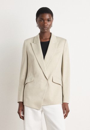 Blazer beige su misura con revers accentuati, due tasche frontali e un tessuto texturizzato. Indossato sopra un top nero, abbinato a pantaloni bianchi.