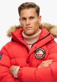 Rød pufferjakke med hætte i pels, sorte lynlåse og broderet patch "Superdry Everest Expedition Co.". Har en ribbet beige sweater under.