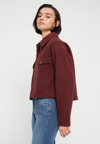 Κοντό μπουφάν μωβ με γιακά, φερμουάρ μπροστά και μία τσέπη στο στήθος, συνδυασμένο με ανοιχτόχρωμο τζιν παντελόνι με διακοσμητικό ράψιμο.