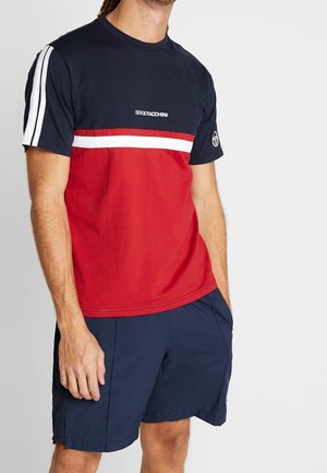 Mężczyzna ubrany w granatowo-czerwoną koszulkę Sergio Tacchini z białymi paskami na rękawach oraz granatowe spodenki, stojący na jednolitym tle.