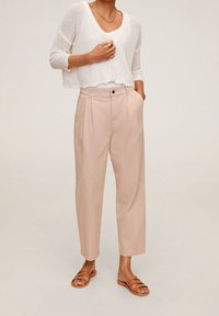 Kobieta w beżowych wysokich spodni z plisami, białym dzianinowym kardiganie do pępka, z warstwowym naszyjnikiem, złotym pierścionkiem i brązowych płaskich sandałach na podwójnych paskach.