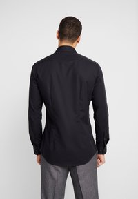 Man som har på sig en åtsittande svart långärmad skjorta och grå byxor, står med ryggen mot kameran framför en ensam bakgrund.