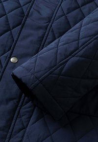 Marineblå quiltet jakke med diamantmønster, en glat tekstur og en metaltrykknapdetalje foran.