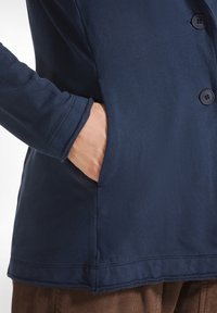 Námořnická modrá bavlněná bunda s knoflíkovou přední částí a boční kapsou, která má hladkou texturu a jednoduchý, čistý design.