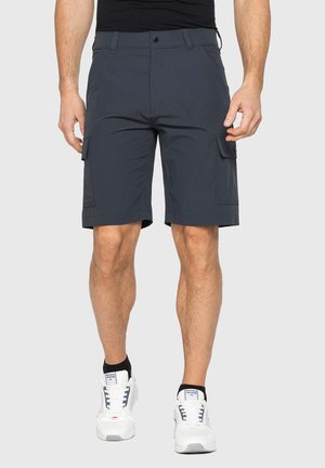 Uomo che indossa pantaloncini cargo grigio scuro con tasche a patta frontali e laterali, sneakers bianche e camicia nera su sfondo uniforme.