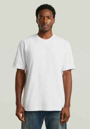 Uomo in piedi di fronte, indossa una maglietta bianca a maniche corte semplice e jeans blu su uno sfondo grigio chiaro.