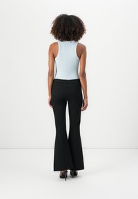 Femme aux cheveux bouclés portant un débardeur bleu clair et un pantalon noir évasé, debout face à un mur blanc.