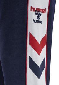 Marineblaue Sweatpants mit weißen Seitenpanels, die mit roten und marineblauen Chevrons sowie dem "hummel"-Logo in roter, strukturierter Stickerei versehen sind. Weiches Material, tapered Fit.