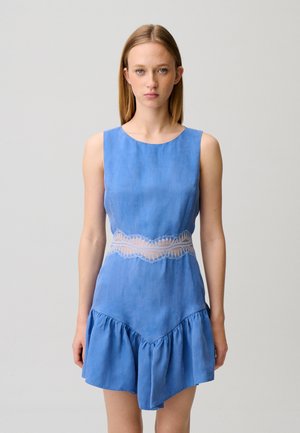 Giovane donna in piedi, indossa un abito blu senza maniche con dettaglio in pizzo alla vita e orlo asimmetrico arricciato, su sfondo chiaro uniforme.