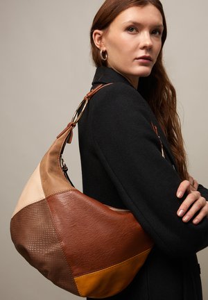 Kvinde med langt brunt hår iført en sort frakke bærer en stor skuldertaske i patchwork-læder i brune, tan og sennepsfarvede nuancer.