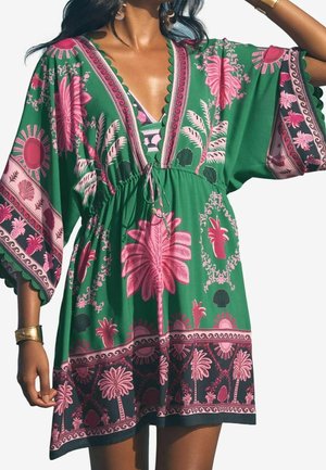 Robe verte à motifs floraux tropicaux roses et palmiers, col en V, manches larges et taille cintrée portée par une personne avec des bijoux en or.