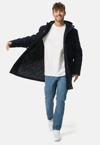 Donkerblauwe herenmantel met een doorgestikte voering aan de binnenkant, knopen aan de voorkant en een capuchon, gecombineerd met lichtblauwe spijkerbroek en witte sneakers.