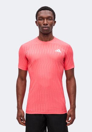 Mann, der frontal steht, trägt ein korallenfarbenes, kurzärmeliges Sportshirt mit weißem Logo auf der Brust und schwarze Shorts.