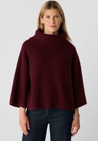 Woman with long blonde hair wearing gold hoop earrings, a loose maroon ribbed turtleneck sweater, and dark fitted pants.