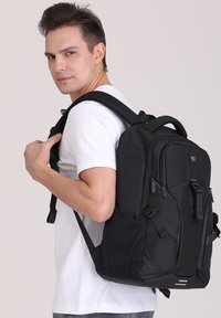 Sac à dos noir en tissu durable, avec plusieurs compartiments, des bretelles rembourrées et des fermetures éclair. Texture lisse avec des accents matériels minimalistes.