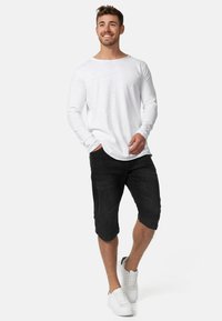 Bijela košulja dugih rukava s okruglim izrezom, u kombinaciji s crnim šortsem do koljena i bijelim tenisicama. Glatka tkanina i ležeran kroj.