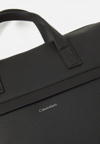 Sac fourre-tout en cuir noir avec une texture granuleuse, un design épuré et un logo "Calvin Klein" en relief sur le panneau avant.