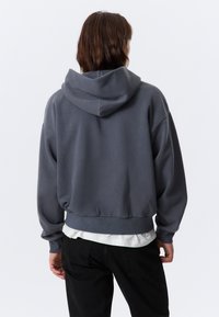 Grå kortermet hettegenser med stor hette, løs passform og ribbestrikket nederkant. Båret over en hvit skjorte, kombinert med svarte bukser.