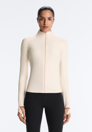 Žena nosiaca béžový dlhý rukáv na zips s otvormi na palce a čierne priliehavé legíny, stojaca pred jednofarebným pozadím.