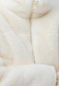 Veste en fausse fourrure crème avec un col haut, une fermeture éclair élégante et une texture matelassée, conçue pour la chaleur et le confort.