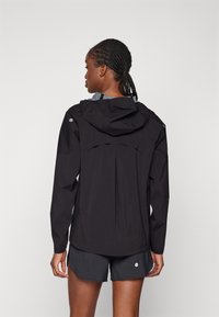 Svart lett jakke med hette, med baklufting og et stilrent design. Kombinert med mørke shorts og glatt tekstur.