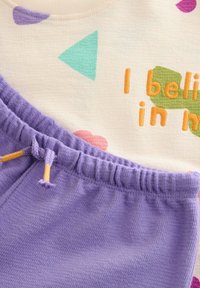 T-shirt i bomull med flerfärgade geometriska mönster och broderad text, kombinerad med mjuka lila handduksshorts med elastisk midja.