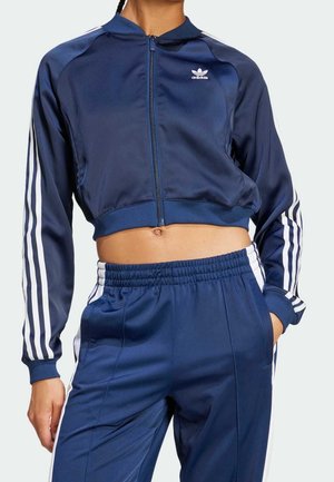 Mujer con chándal azul marino de Adidas con rayas blancas en las mangas y pantalones, con chaqueta corta y pantalones con cintura elástica.
