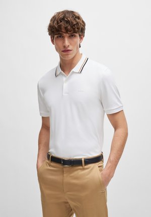 Weißes Poloshirt in Slim-Fit, mit einem schwarz-goldenen gestreiften Kragen. Getragen mit beigefarbenen Hosen und einem schwarzen Gürtel, was lässige Eleganz betont.