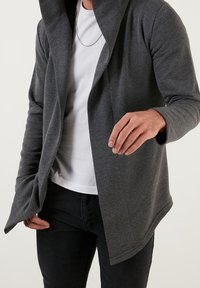 Kolsvar färg, långärmad jacka med huva, med en draperad framsida. Tillsammans med en vit t-shirt och svarta jeans. Slät textur.