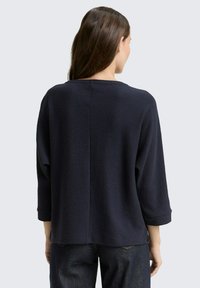 Donkerblauwe pullover met een textuur, geribbelpatroon. Heeft driekwart mouwen en een losse pasvorm, met naaddetails op de rug.