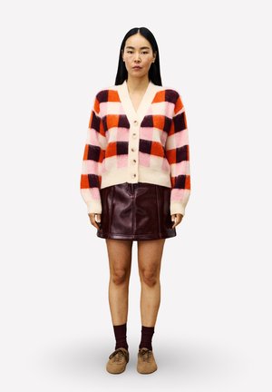 Femme portant un cardigan à carreaux multicolore avec des boutons beiges, une jupe en cuir bordeaux foncé, des chaussettes sombres et des chaussures marron, se tenant devant un fond blanc.