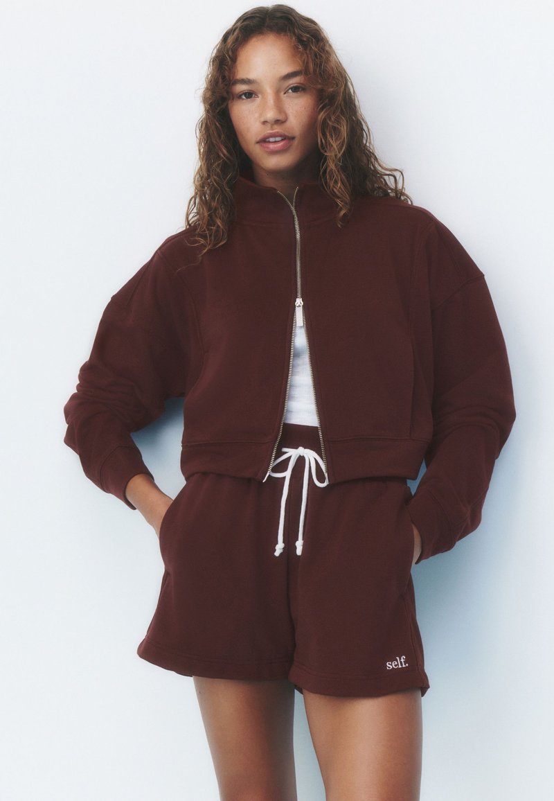 Woman with curly hair wears a dark brown zip-up sweatshirt and matching shorts with white drawstring and "self." logo on left leg.
