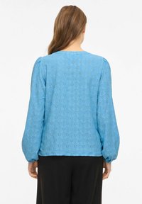 Hele sinine pluus pikkade, paeltega varrukatega, millel on tekstuuriga silmaaugumuster ja ümmargune kaelus. Kanditud koos kontrastsete mustade pükstega.